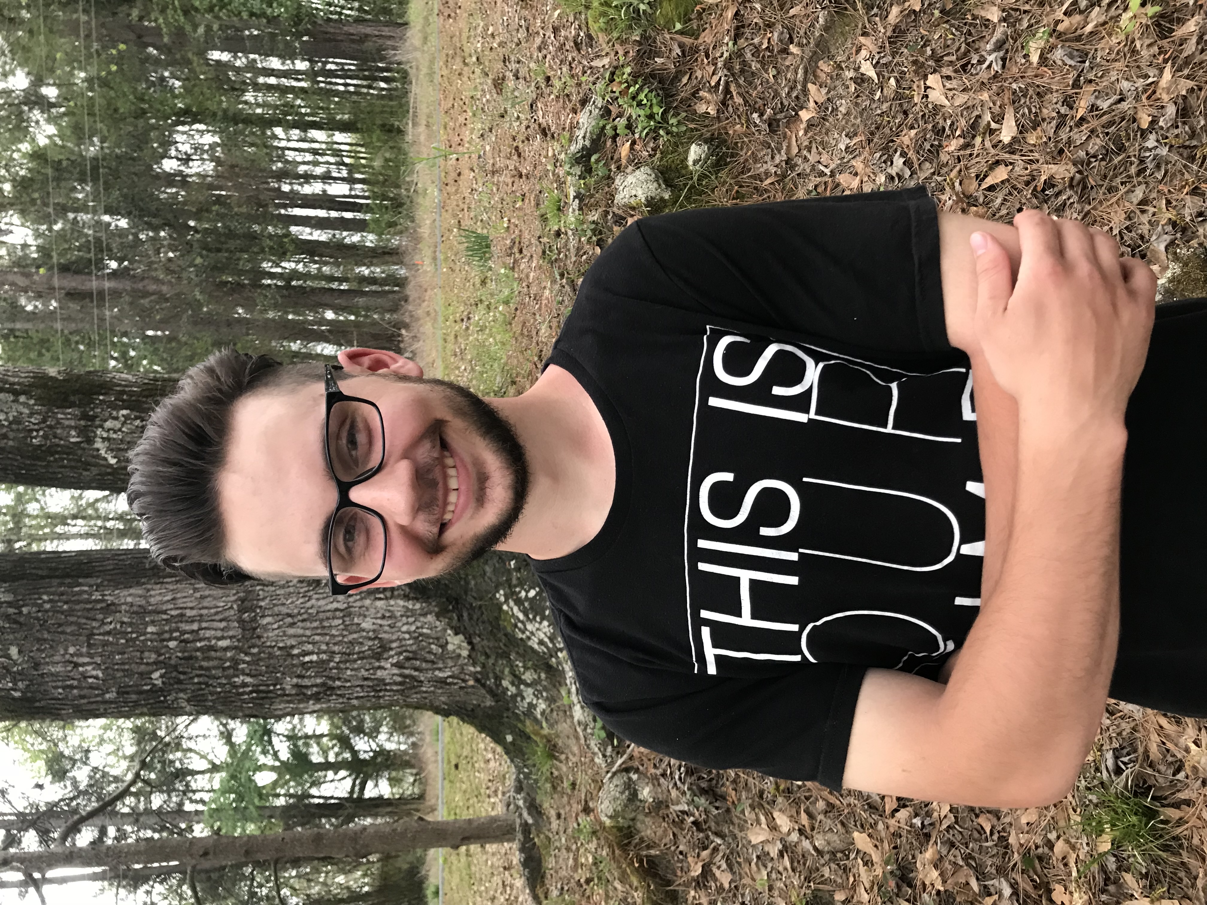 young man wearing glasses smiling into the camera with a hill and trees behind him