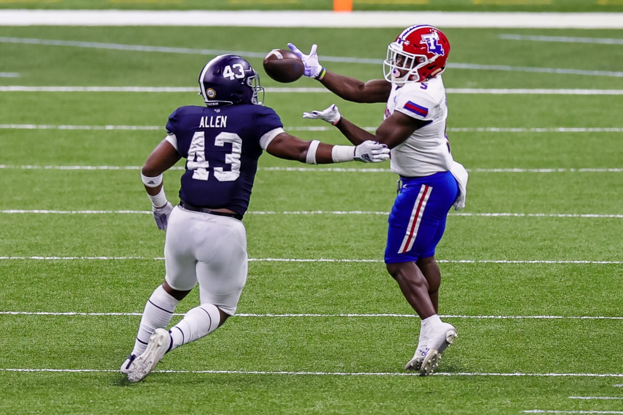 a LA tech football player with a white, red helmet, and blue pants uniform
             catching a football with an opposing team player in a blue and white uniform trying to interfere with the catch
             in the middle of a daytime game